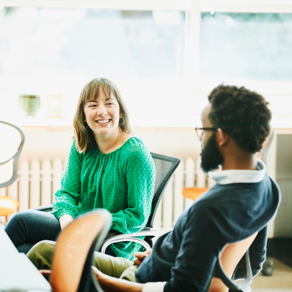 A smiling businesswoman engaged in a discussion with her colleague at a workstation in a modern office. Both are focused and exchanging ideas, surrounded by computers, documents, and office supplies. The atmosphere is collaborative and professional, reflecting a productive work environment.